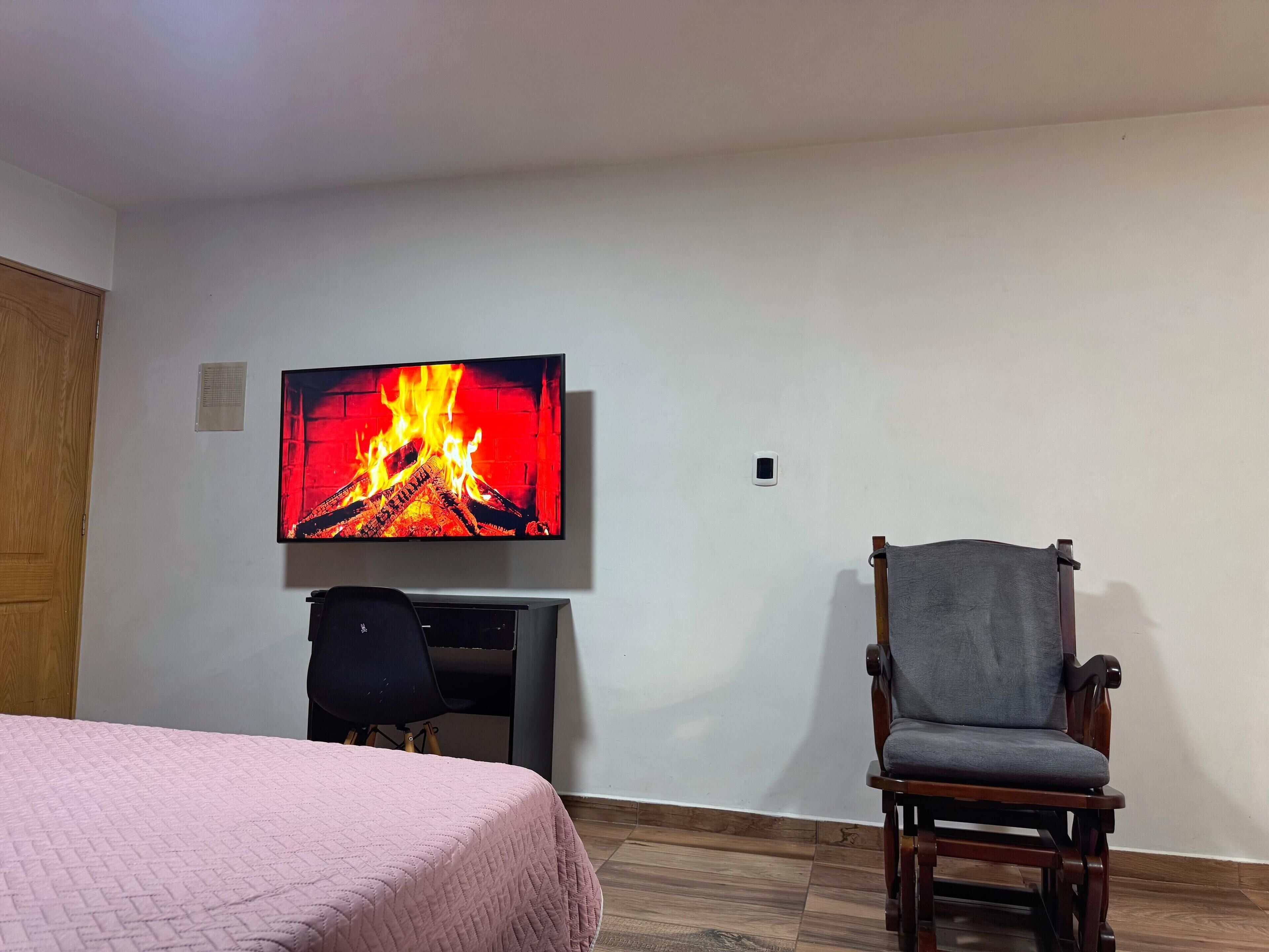 Una habitación con una cama, un escritorio, una silla y un televisor que muestra una escena de chimenea.