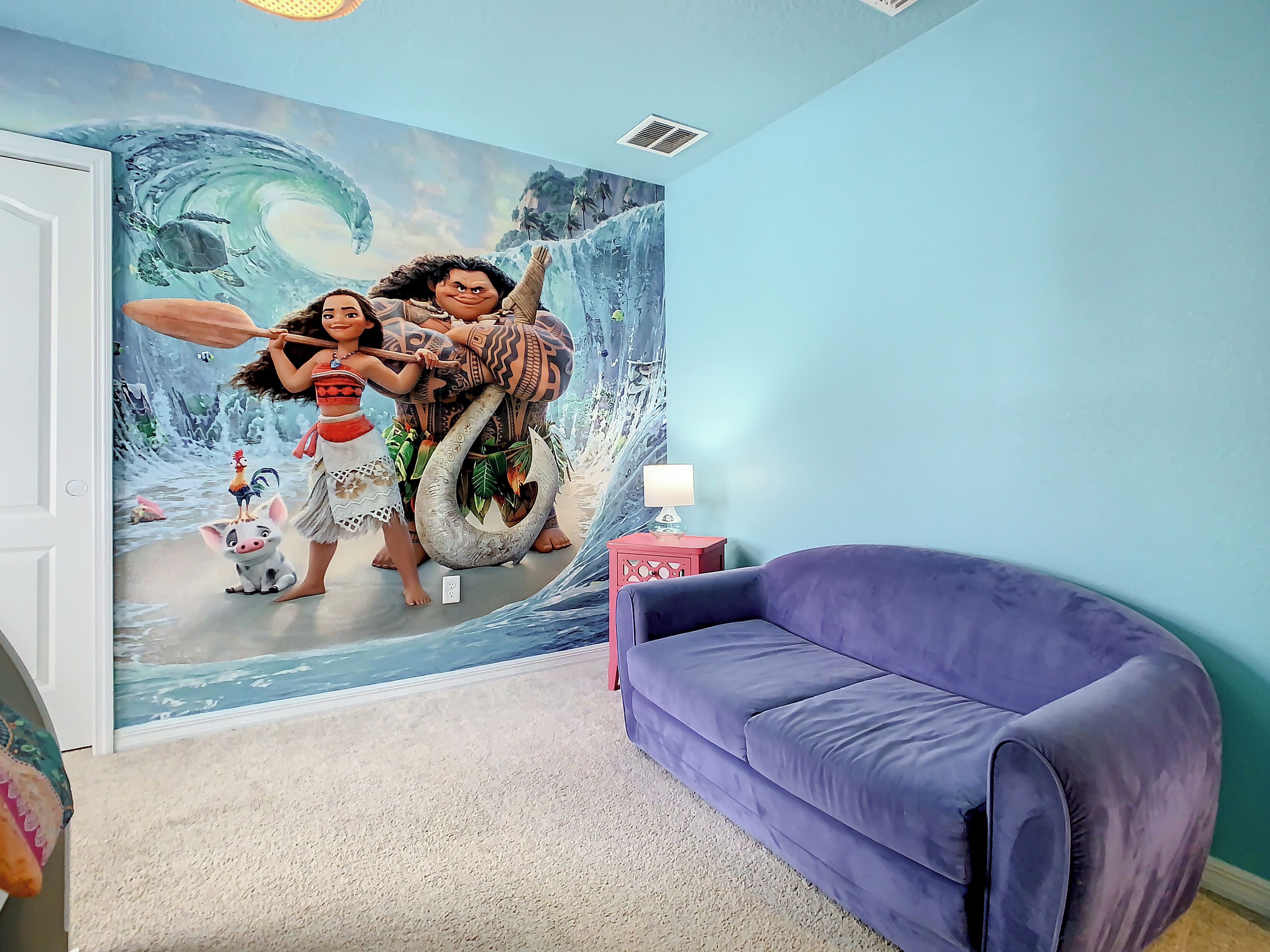 Habitación con un mural de paisaje tropical, un sofá morado y una mesita con una lámpara.