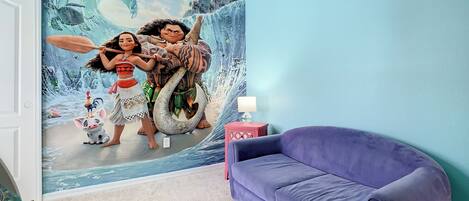 Habitación con un mural de paisaje tropical, un sofá morado y una mesita con una lámpara.