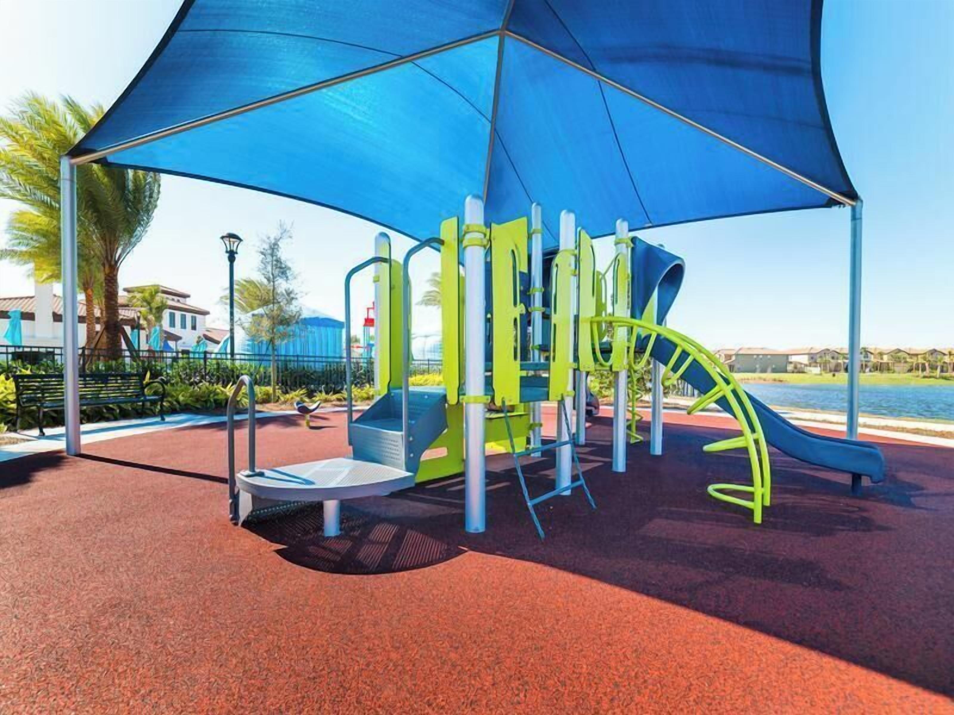 An outdoor playground with a slide and climbing structures, surrounded by a red rubberized surface and a blue canopy.