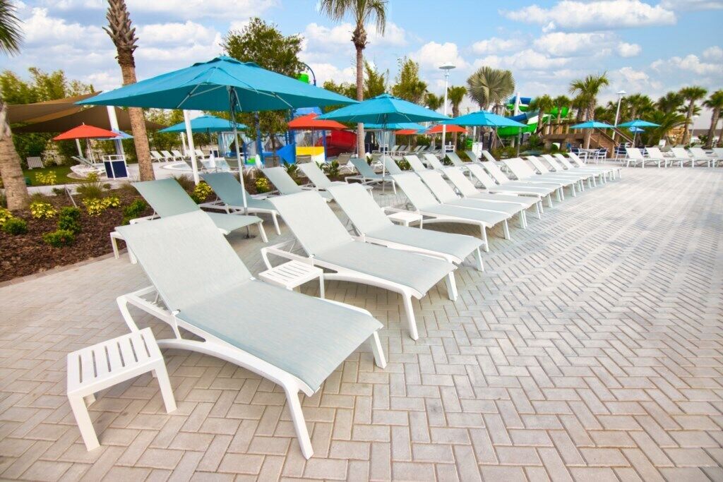 A row of white lounge chairs with blue cushions, arranged on a paved walkway with palm trees and umbrellas in the background.