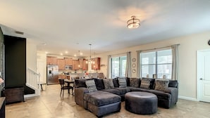 A spacious living room with a sectional sofa and ottoman, a dining area, and a kitchen in the background.