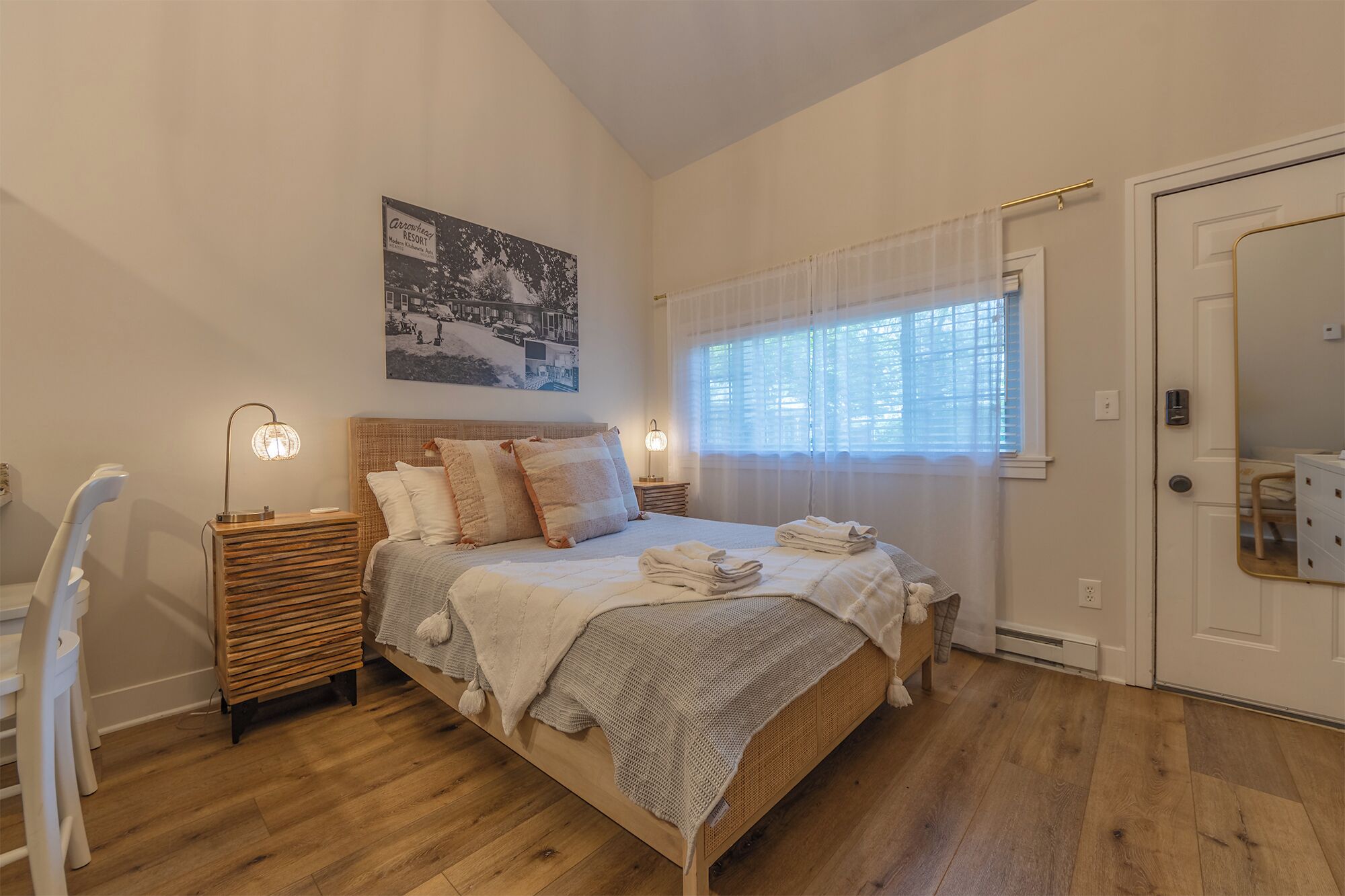 A bedroom with a bed, bedside tables, a lamp, a window with curtains, a mirror, and a wall-mounted picture.