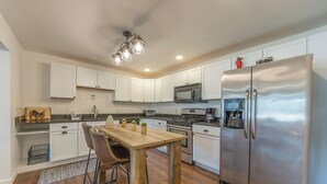 A modern kitchen with white cabinets, stainless steel appliances, a wooden dining table, and a black metal chair. - Timber Lodge Walkable to shops, shoreline & craft beer (Douglas)