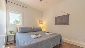 A bedroom with a large bed, a bedside table with a lamp, a small plant, and a window with a view of trees. - Timber Lodge Walkable to shops, shoreline & craft beer (Douglas)