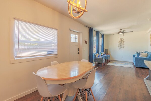 A dining area with a round table and chairs, a ceiling fan, and a living room with a sofa and a window with blinds.