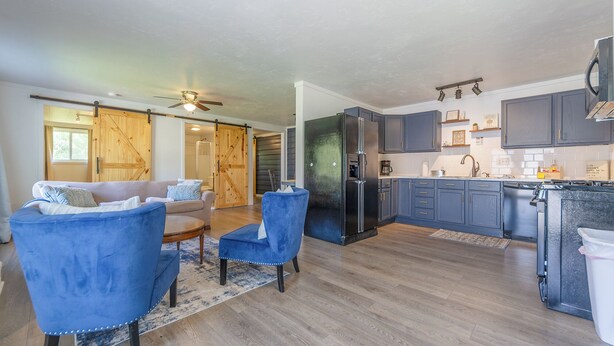 A spacious living area with a sofa and two blue armchairs, a kitchen with dark blue cabinets and a black refrigerator, and a sliding barn-style door leading to another room. - Hickory Cottage: Cozy 2-Bedroom in Douglas, MI, close to beaches, downtown, antiques and more!* (Douglas)