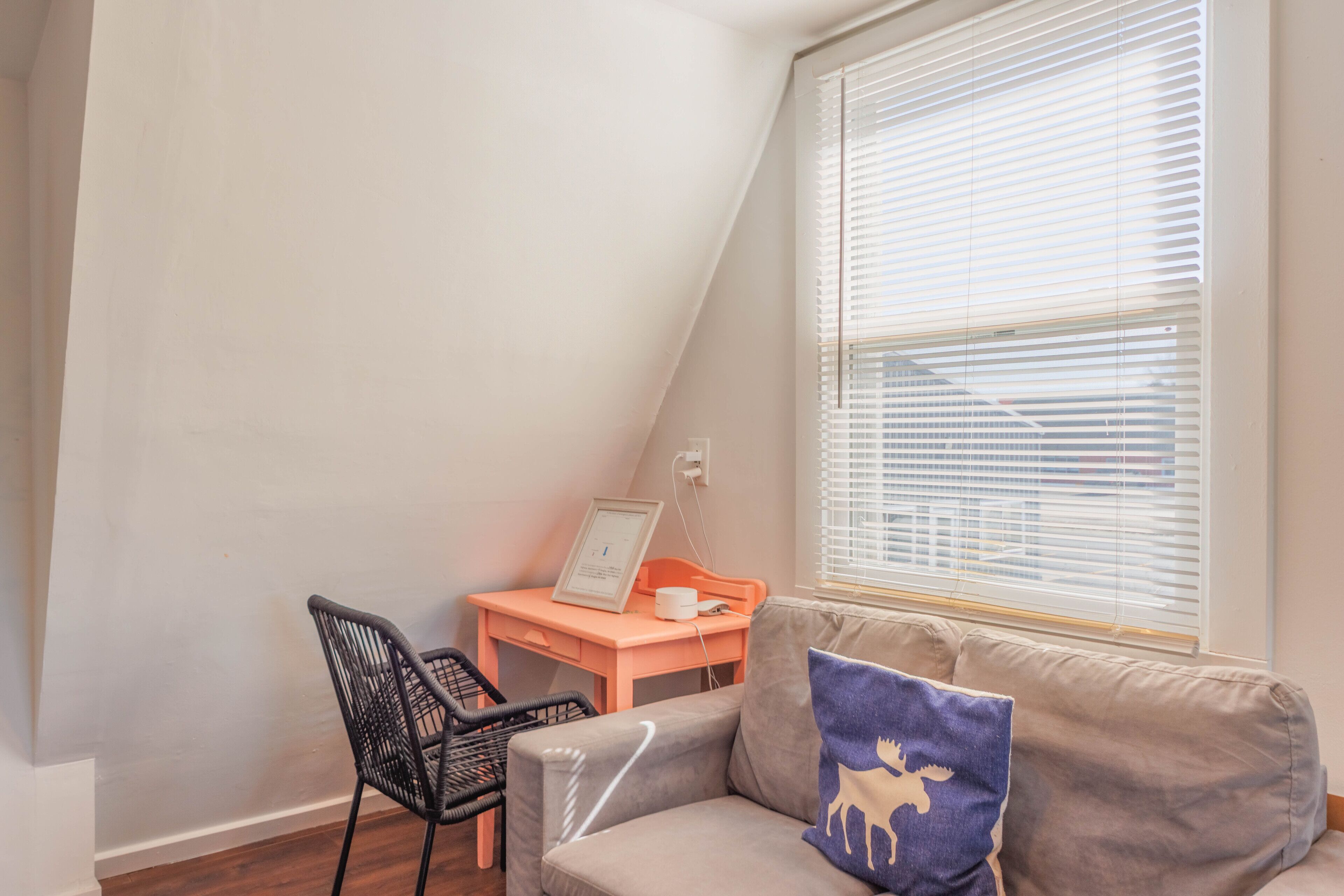 Una habitación con techo inclinado, una ventana con persianas, un escritorio con silla y un sofá con cojín.