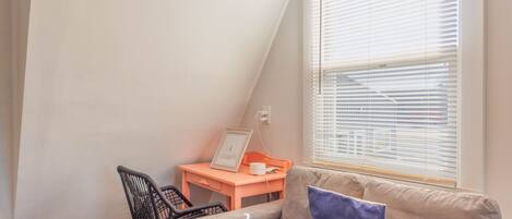 Una habitación con techo inclinado, una ventana con persianas, un escritorio con silla y un sofá con cojín.