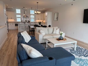 A modern living room with a sectional sofa, a coffee table, and a dining area with a white table and chairs. - Ocean Breeze 102: Dog Friendly, Brand New Townhome, Close to Beach! (Kill Devil Hills)