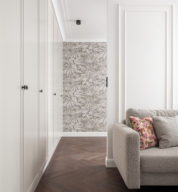 A room with a herringbone-patterned wooden floor, a grey upholstered sofa with a floral pillow, and a white door with a small black handle.