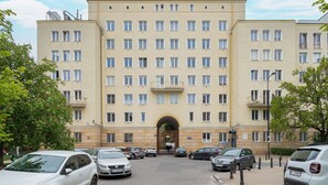 Un edificio de varios pisos con una entrada central, varias ventanas y coches aparcados en la parte delantera.