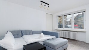 A modern living room with a grey sofa, a white bed, and a black side table. A large window offers a view of buildings outside.