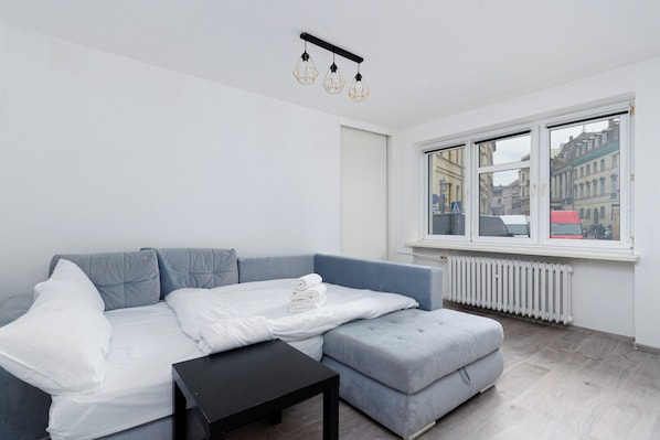 A modern living room with a grey sofa, a white bed, and a black side table. A large window offers a view of buildings outside.