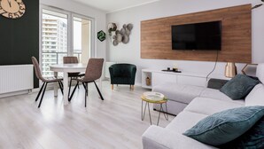 A modern living room with a grey sofa, a dining table with chairs, a TV mounted on a wooden panel, and a large clock on the wall.