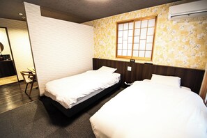 A hotel room with two single beds, a wooden headboard, a floral wallpaper, a window with a grid pattern, and a desk with a chair.