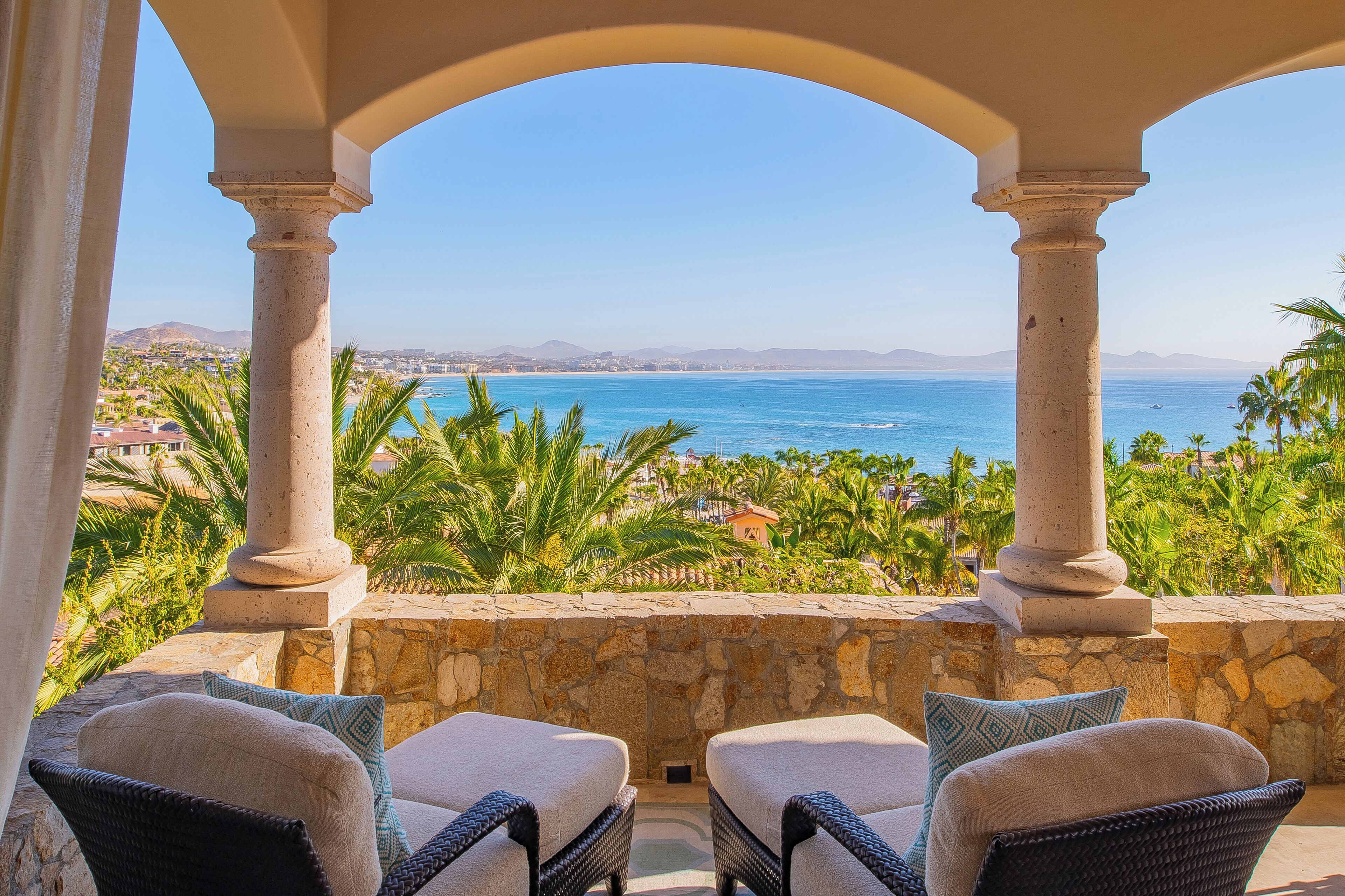 Un balcon avec deux fauteuils en osier et un mur de pierre, offrant une vue sur la côte, avec des palmiers et un ciel bleu dégagé.