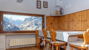 Zona pranzo con un'ampia finestra che si affaccia su un paesaggio montuoso, sedie di legno con cuscini bianchi e un mobiletto in legno con ripiano bianco.