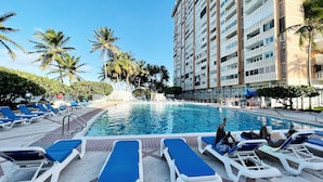 A hotel pool area with lounge chairs, palm trees, and a tall building in the background.