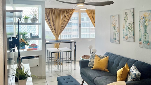 A modern living room with a grey sofa, yellow cushions, a dining table, and a ceiling fan. There are potted plants and artwork on the walls.