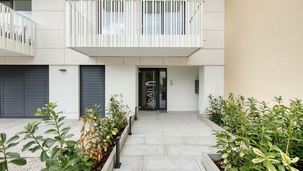 Un edificio moderno con techo plano, un balcón con barandas blancas y una puerta con panel de vidrio.