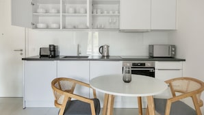 Una cocina moderna con armarios blancos, una mesa redonda para comer, dos sillas de mimbre, una cafetera, un hervidor, un tostador y un lavavajillas.