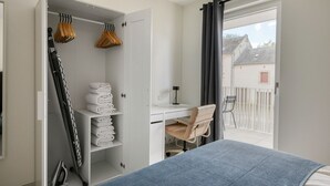 Un dormitorio con cama, escritorio y silla. Se observa un armario con ropa colgada y una estantería con toallas dobladas. Se ve una ventana con cortina y un balcón con una silla en el exterior.