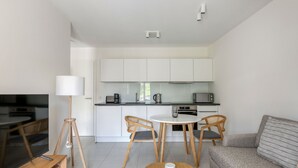 Una sala de estar moderna con televisor, sofá y una mesa de centro. Se observa un comedor con una mesa y sillas. Se ve la cocina, con gabinetes y electrodomésticos blancos.