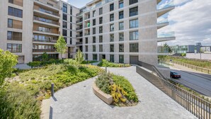 Un moderno edificio residencial de varios pisos con balcones, un patio ajardinado y una calle con un automóvil pasando.