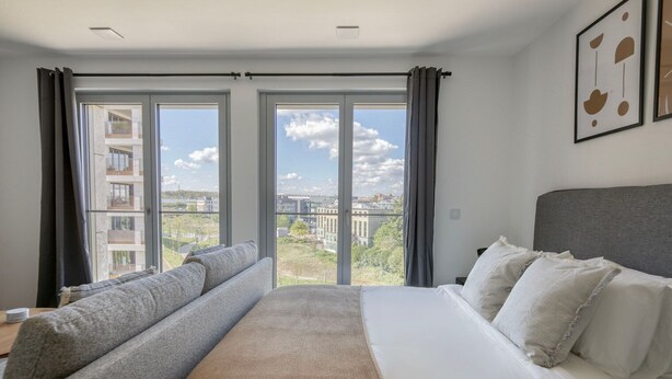 Una habitación de hotel moderna con una cama grande, un sofá y vistas de un paisaje urbano a través de ventanas de piso a techo.