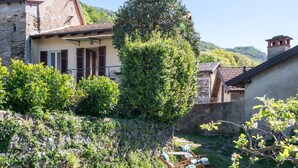 Una casa de piedra con techo de tejas, rodeada de abundante vegetación y un muro de piedra.
