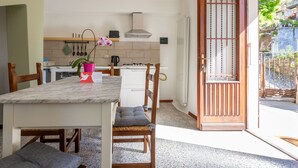 Una cocina moderna con armarios blancos, una estufa y un lavamanos. Se observa una mesa de comedor con sillas y un florero. Se ve una puerta de madera con una reja metálica.