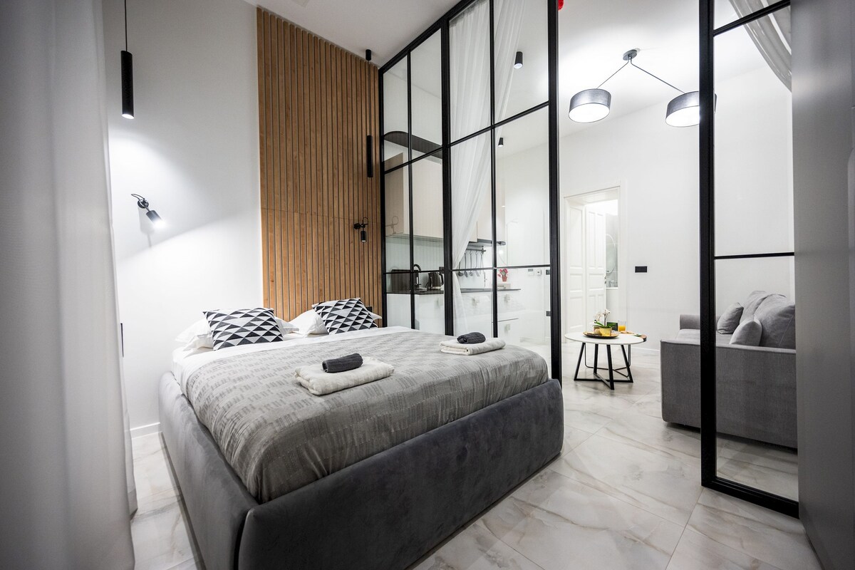 A modern bedroom with a large bed, a wooden panel wall, a glass door leading to a balcony, and a grey sofa.