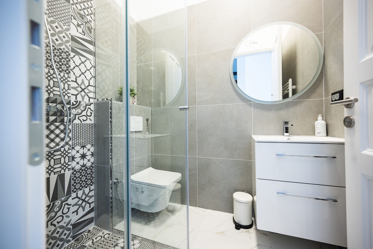 A modern bathroom with a glass shower enclosure, a toilet, a sink with a mirror, and a trash bin.