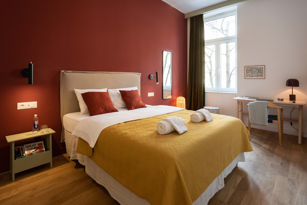 Una habitación de hotel con cama, escritorio, silla, ventana y mesita de noche con lámpara.
