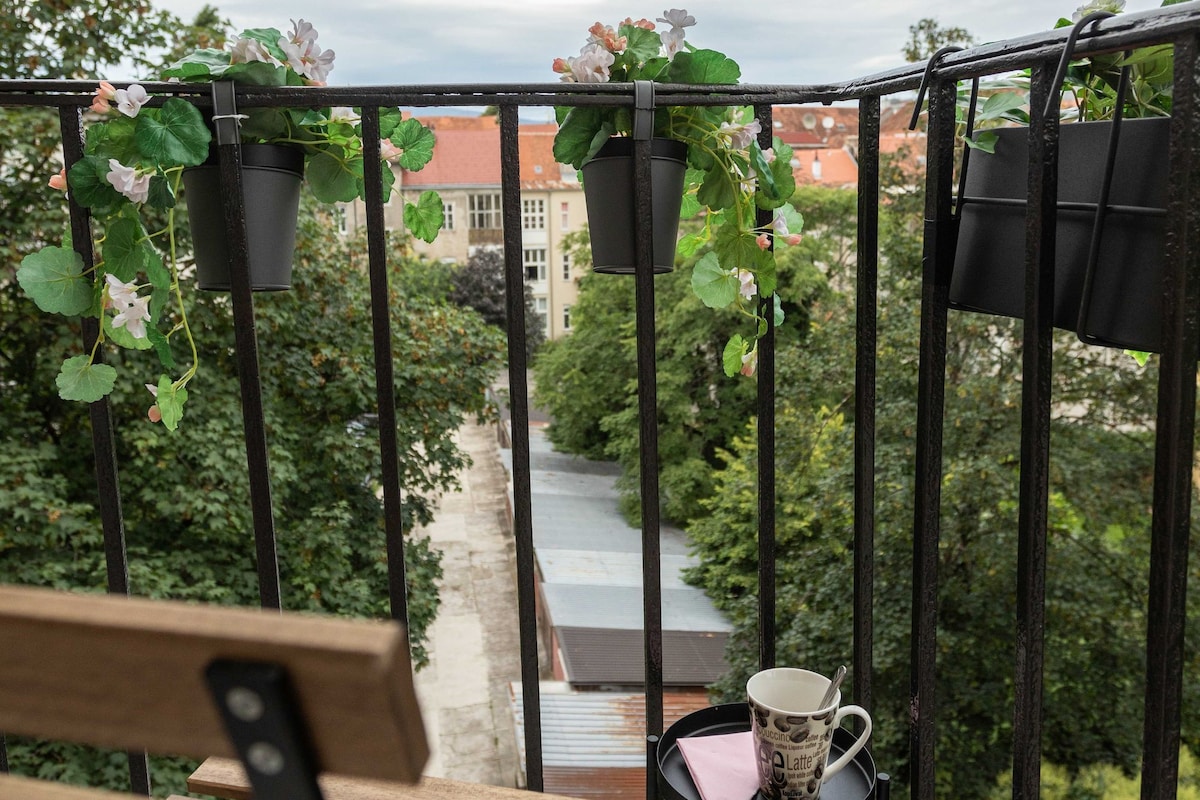 Un balcón con banca de madera, una mesita con taza y platillo, y macetas colgantes con plantas verdes.