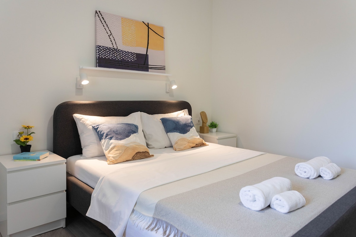 Una cama bien hecha con almohadas, una mesita de noche con un libro y una planta en maceta, y una obra de arte colgada en la pared sobre la cama.