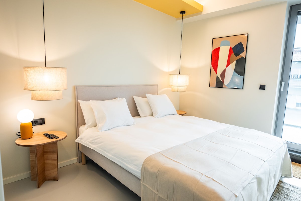 A hotel room with a large bed, bedside lamps, a bedside table with a lamp, a remote control, and a wall-mounted abstract painting.