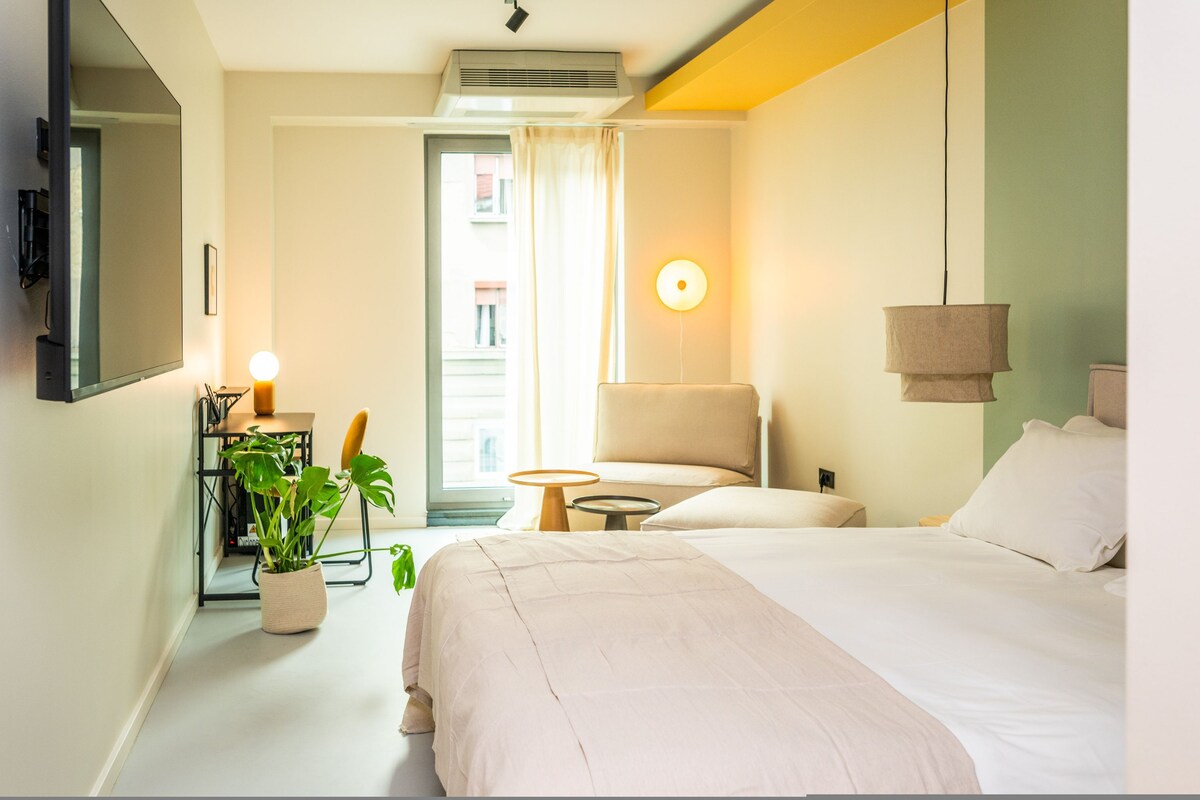 A hotel room with a bed, a chair, a small table, a potted plant, and a wall-mounted lamp.