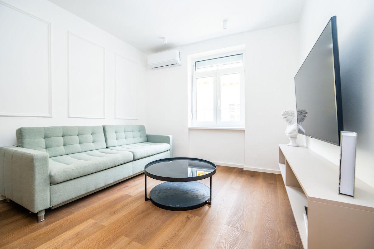 A modern living room with a sofa, a round coffee table, a flat-screen TV, and a window with natural light.