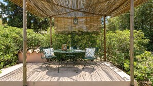 Uma área de estar coberta ao ar livre, com mesa e cadeiras, cercada por vegetação.