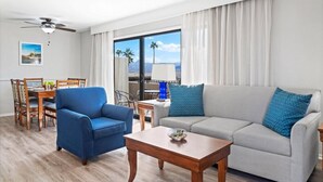 Een moderne woonkamer met een bank, fauteuil, eettafel en stoelen. Door grote ramen is een balkon zichtbaar met uitzicht op palmbomen en bergen.