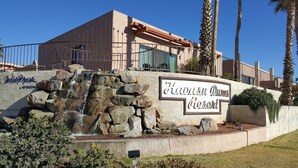 A building with a sign reading 'Hawaiian Dunes Resort,' a stone wall with a waterfall feature, and palm trees. - Lake Havasu City | Couples Lake Retreat | Pool + Hot Tub (Lake Havasu)