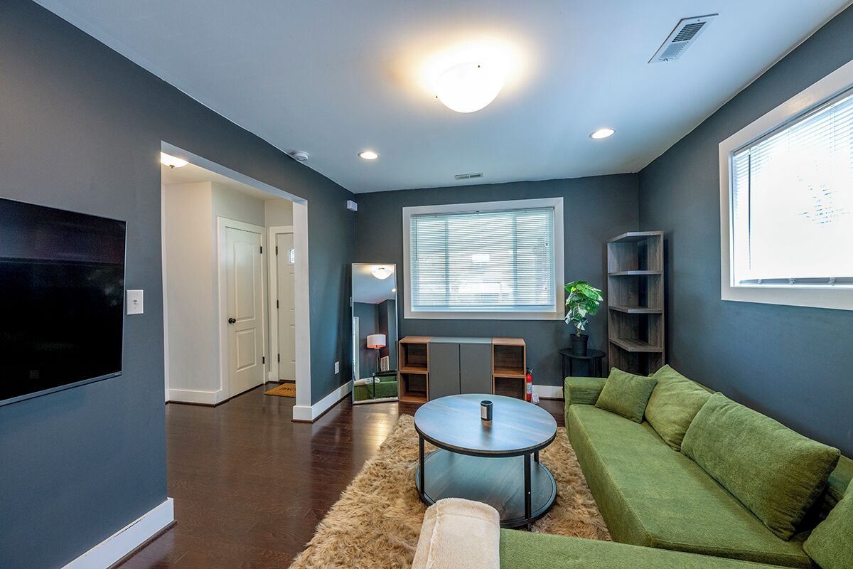 A modern living room with a green sofa, a round coffee table, and a TV mounted on the wall. There is a window with blinds, a radiator, and a small plant on a side table.