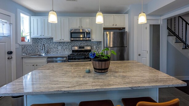 A modern kitchen with white cabinets, stainless steel appliances, a marble island, pendant lights, and a potted plant.