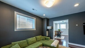 A modern living room with a green sectional sofa, a round wooden coffee table, and a large window with blinds.