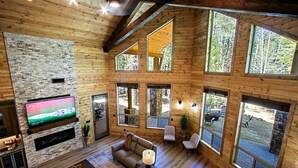 A spacious living area with a stone wall, large windows, a flat-screen TV, and a cozy seating arrangement. - Luxury Cabin in a woods! Private location 7000 ft elevation, 4 seasons! (Prescott)