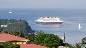 Ein großes Kreuzfahrtschiff liegt im Hafen fest, mit freiem Blick auf die umliegende Landschaft, darunter ein Gebäude mit rotem Dach und üppiger Vegetation.