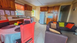 Eine Küche mit Holzausstattung, einem Kühlschrank und einer Mikrowelle. Ein Esstisch mit Stühlen und einer Obstschale steht im Raum. Im Wohnbereich befindet sich eine Couch mit bunten Kissen.