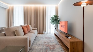 Ein modernes Wohnzimmer mit einer Couch, einem Fernseher auf einem Holzboden, einer Stehlampe und einem großen Fenster mit Vorhängen.